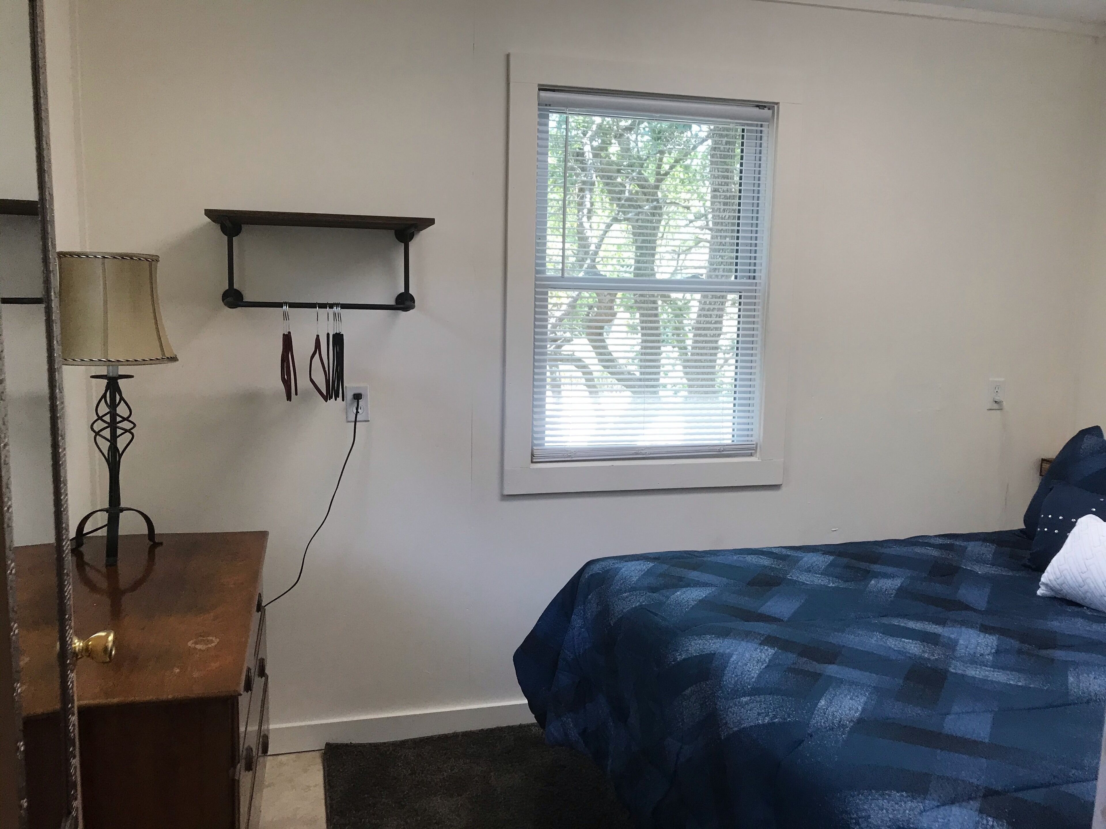 Un dormitorio con cama, mesita de noche, lámpara, ventana con persianas y estante en la pared.