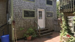 Ein kleines Haus mit einer Haustür, zwei Fenstern, einer Holzterrasse und einem Blumentopf.