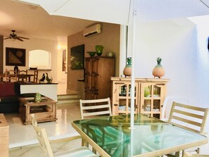 A dining area with a glass-top table, wooden chairs, a white umbrella, and a wooden cabinet with decorative items.