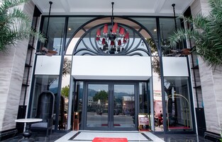 Entrada de hotel con diseño moderno, que incluye un gran ventanal, una araña y una alfombra roja que conduce a la entrada.