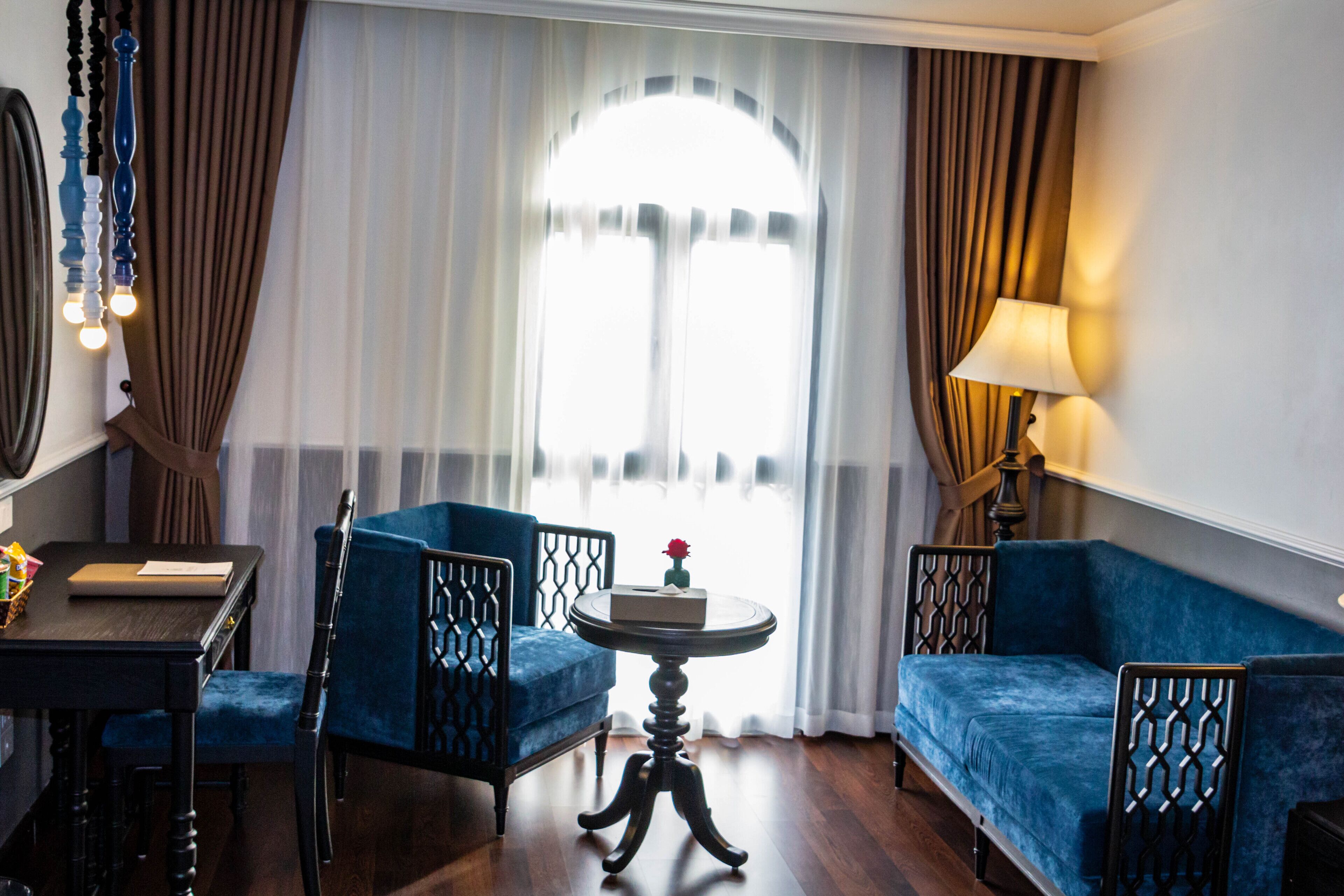 A room with a blue sofa, a round table with a vase of flowers, a chair, a lamp, and a large window with curtains.