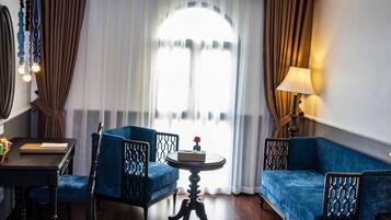 A room with a blue sofa, a round table with a vase of flowers, a chair, a lamp, and a large window with curtains.