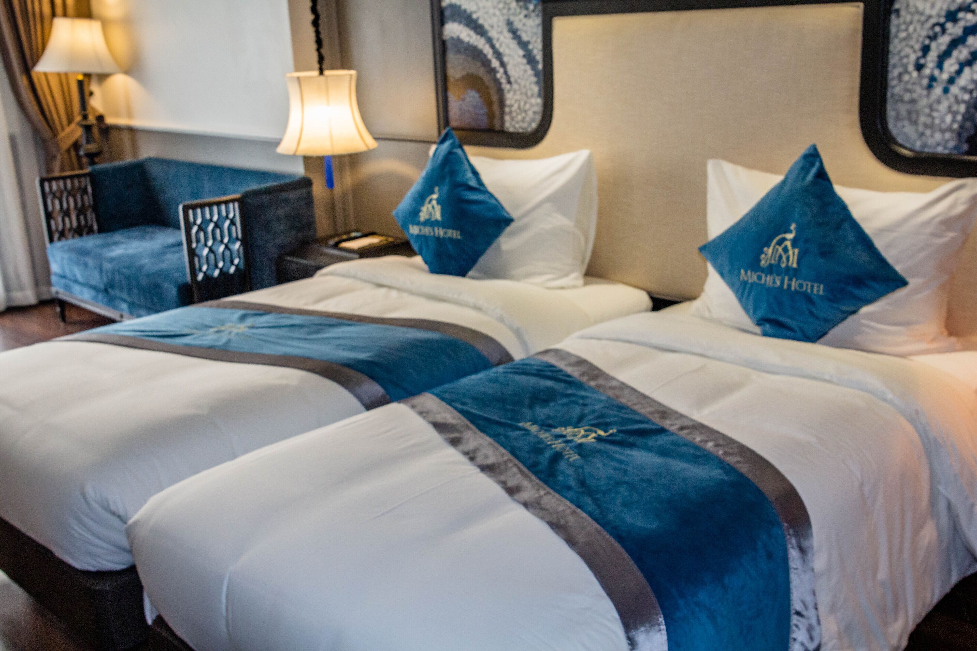 Habitación de hotel con dos camas, cada una con una almohada azul y cabecera blanca. Hay una mesita de noche con una lámpara y un teléfono.