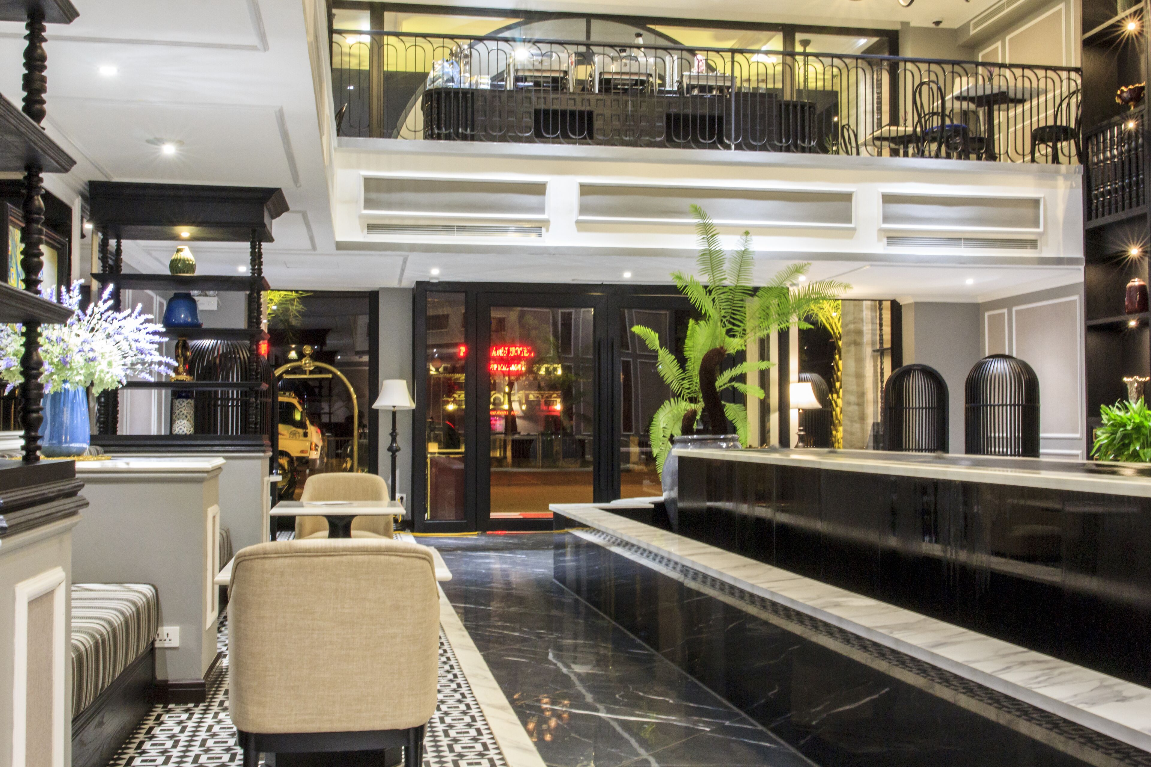 Um hall de hotel com uma recepção de mármore preto, área de estar com cadeiras, escada que leva ao andar superior e uma grande planta.