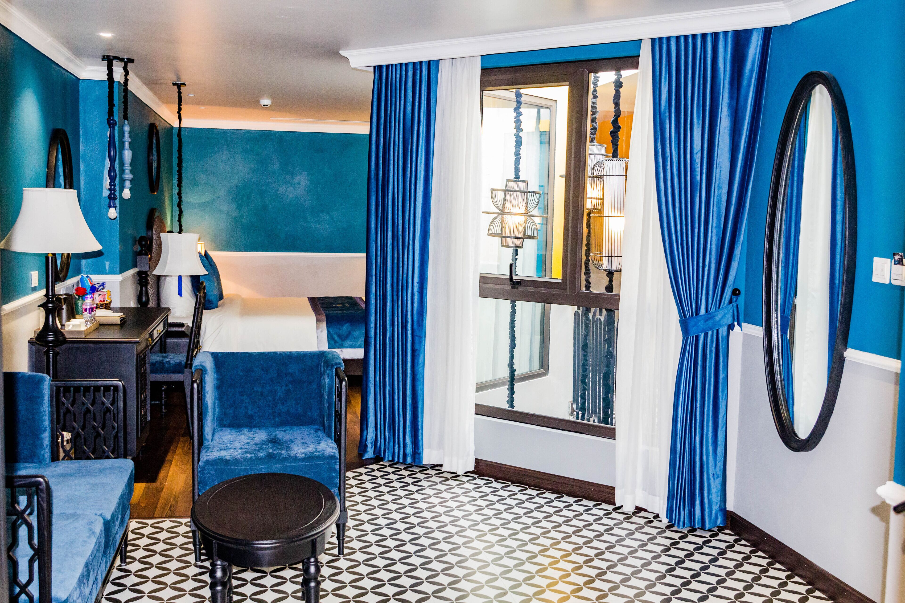 A hotel room with a blue and white color scheme, a bed, a desk with a lamp, a chair, and a patterned floor.