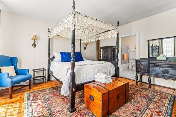 A four-poster canopy bed with a white bedspread and blue pillows, a blue armchair, a wooden chest of drawers, and a patterned rug.
