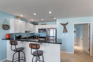 Una cocina con armarios blancos, un refrigerador de acero inoxidable, un microondas y un lavamanos. Hay dos taburetes de bar y un reloj en la pared.
