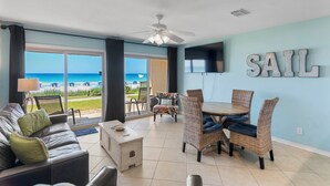 Ein Zimmer mit Blick auf den Strand, einem Deckenventilator, einem Esstisch mit Stühlen und einem Schild mit der Aufschrift 'SAIL' an der Wand.