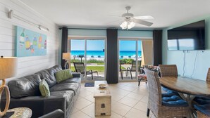 Ein Wohnzimmer mit Blick auf den Strand, einem Deckenventilator, einem Esstisch mit Stühlen und einem Flachbildfernseher.