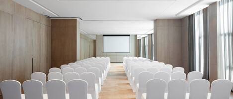 Une salle de conférence avec des rangées de chaises blanches, un grand écran à l’avant et de grandes fenêtres sur un des côtés.