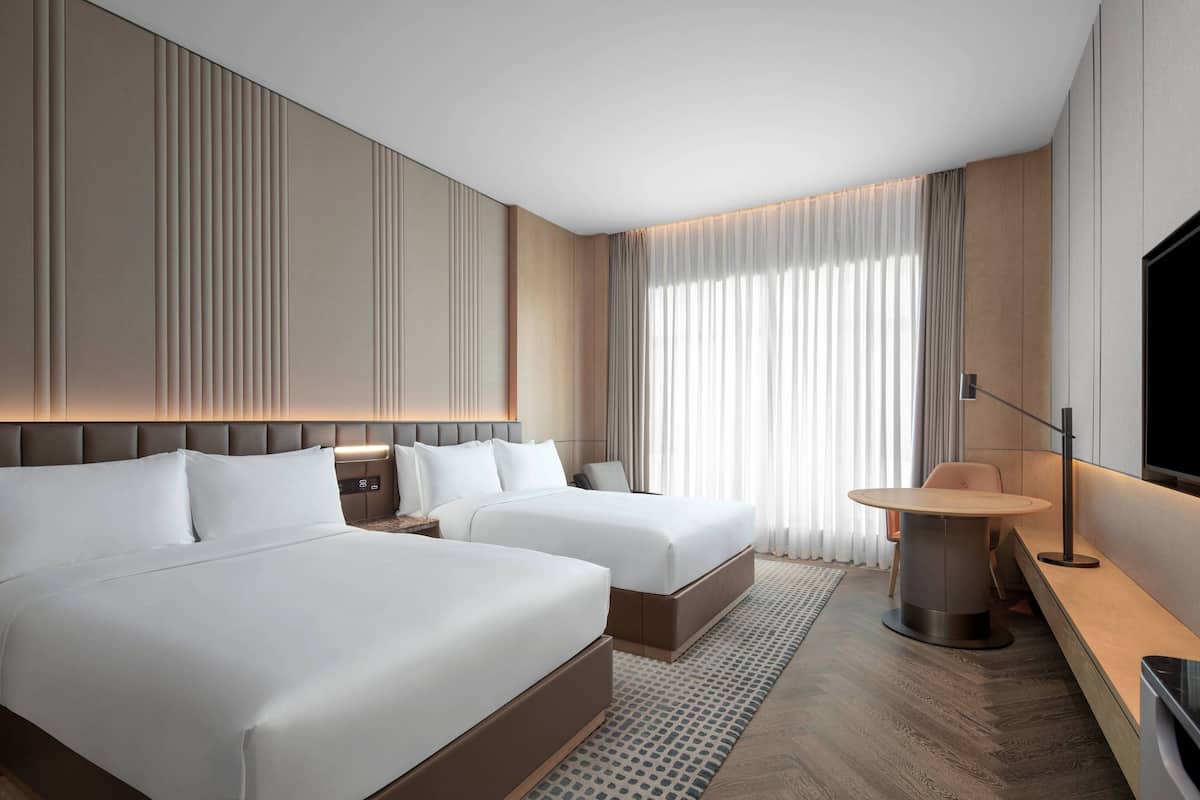 Une chambre d’hôtel moderne avec deux lits, une table à manger ronde, un téléviseur et une grande fenêtre avec des rideaux.