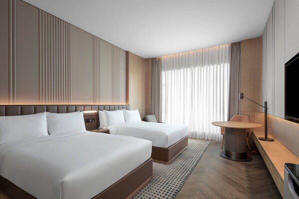 Una habitación de hotel moderna con dos camas, una mesa redonda para comer, un televisor y un amplio ventanal con cortinas.