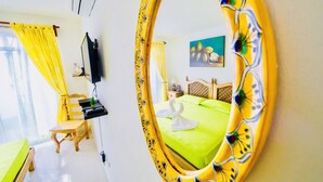 Ein Hotelzimmer mit einem grünen Bett, einem Holzstuhl, einem Schreibtisch, einem Fernseher und einem Spiegel mit floralem Rahmen.