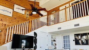 A spacious living room with a high ceiling, wooden beams, a large flat-screen TV, a comfortable sofa, and a staircase with white railings. - Beautiful Beach House with Gulf views, 2 min walk to Beach!!  (Galveston)