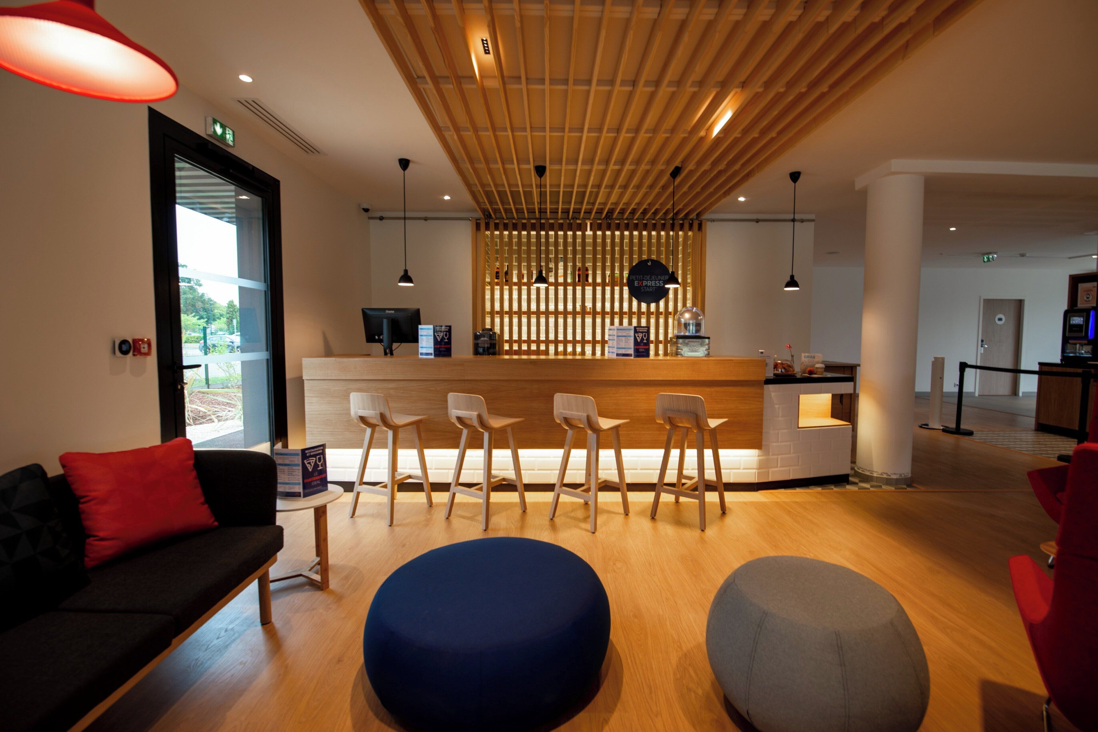 A reception area with a wooden reception desk, bar stools, a blue ottoman, and a black sofa with a red pillow.