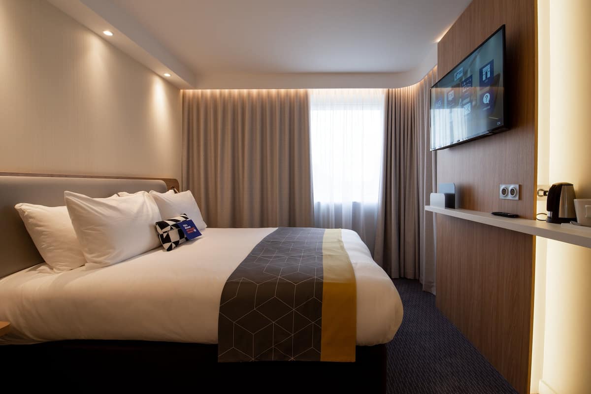 Une chambre d’hôtel avec un grand lit, un téléviseur à écran plat fixé au mur, un bureau avec une machine à café et une fenêtre avec des rideaux.