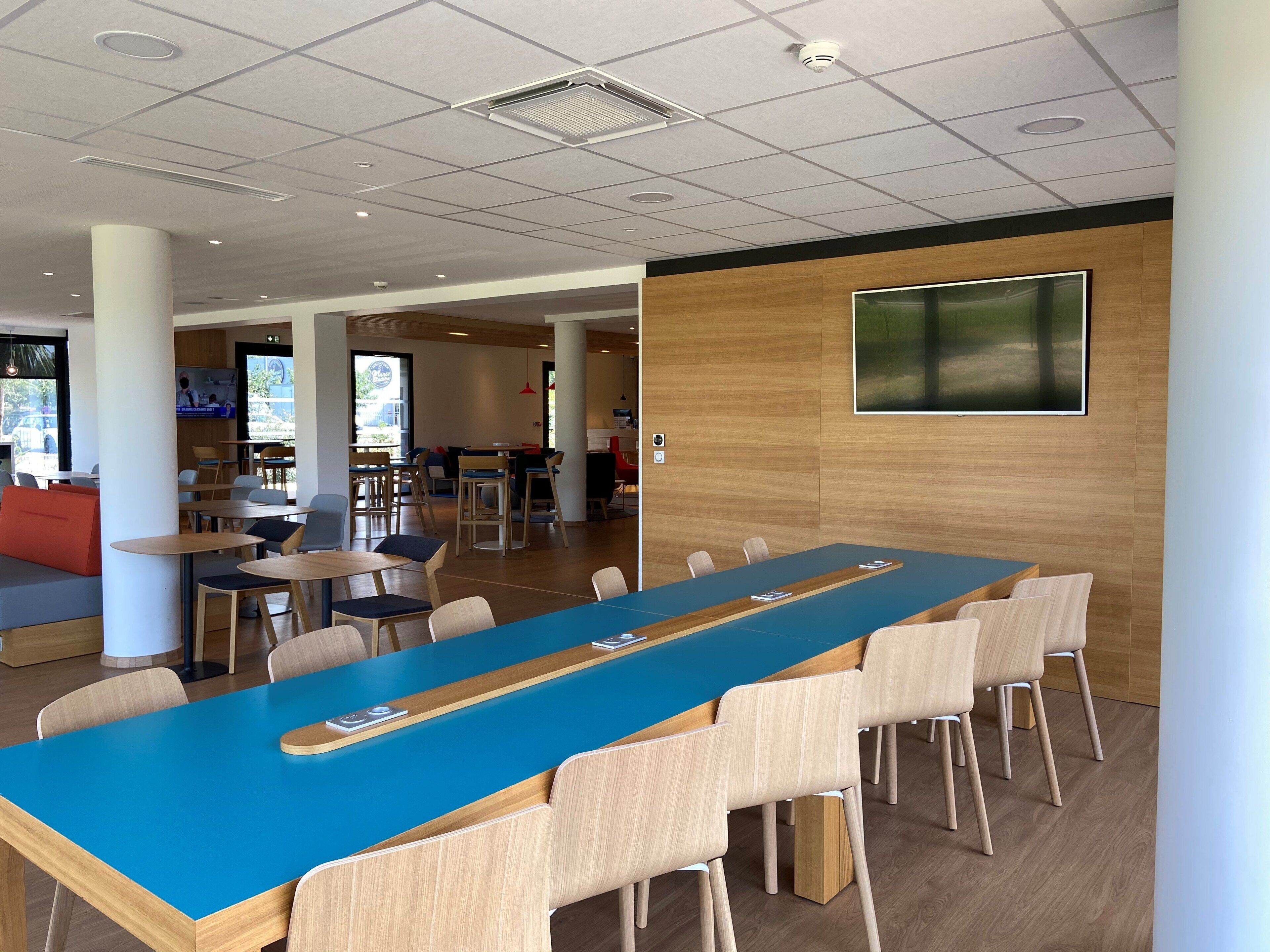 Un espace intérieur moderne avec une longue table bleue, des chaises en bois et un grand écran mural.