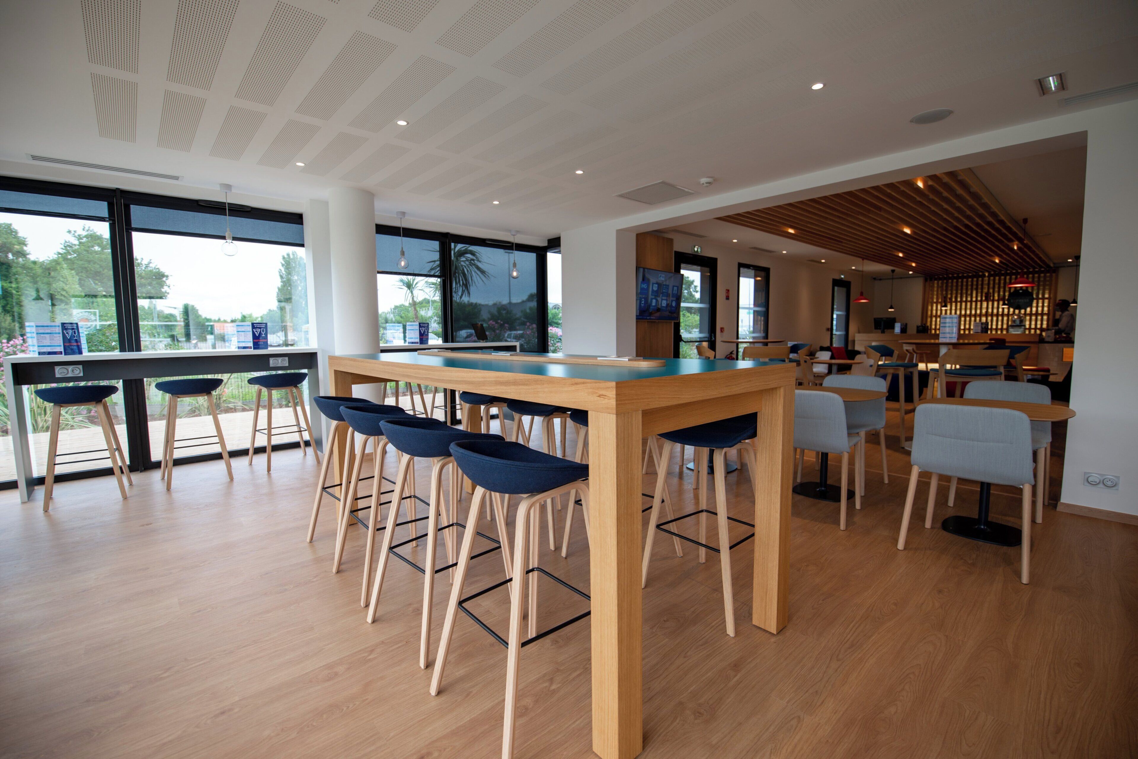 A modern bar area with wooden tables and blue bar stools, large windows, and a view of greenery outside.