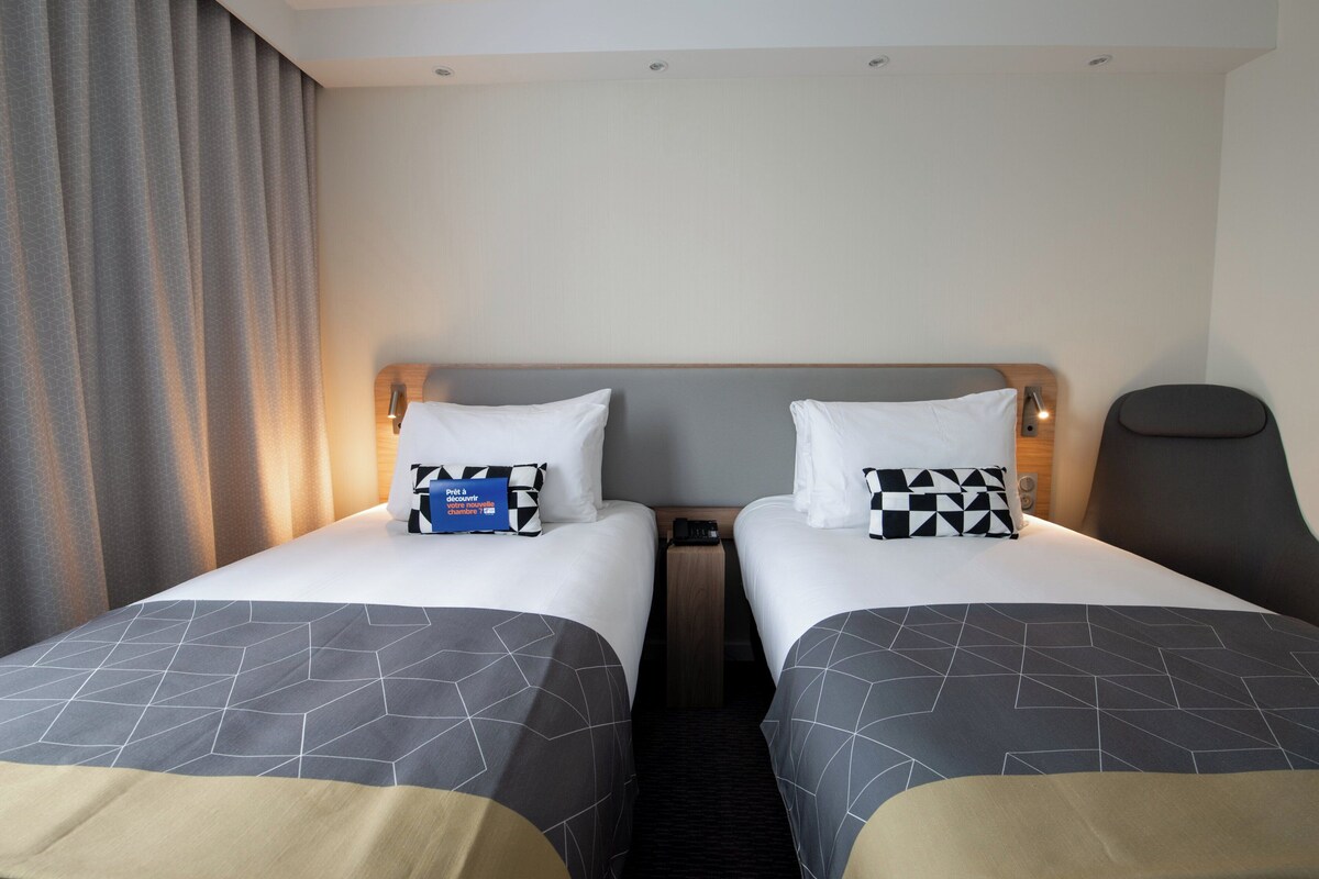 Hotellihuone, jossa on kaksi sänkyä. Jokaisessa sängyssä on geometrisesti kuvioitu peitto, tyyny ja yöpöytä, jossa on puhelin.