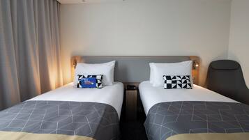 A hotel room with two beds, each with a geometric-patterned bedspread, a pillow, and a bedside table with a phone.