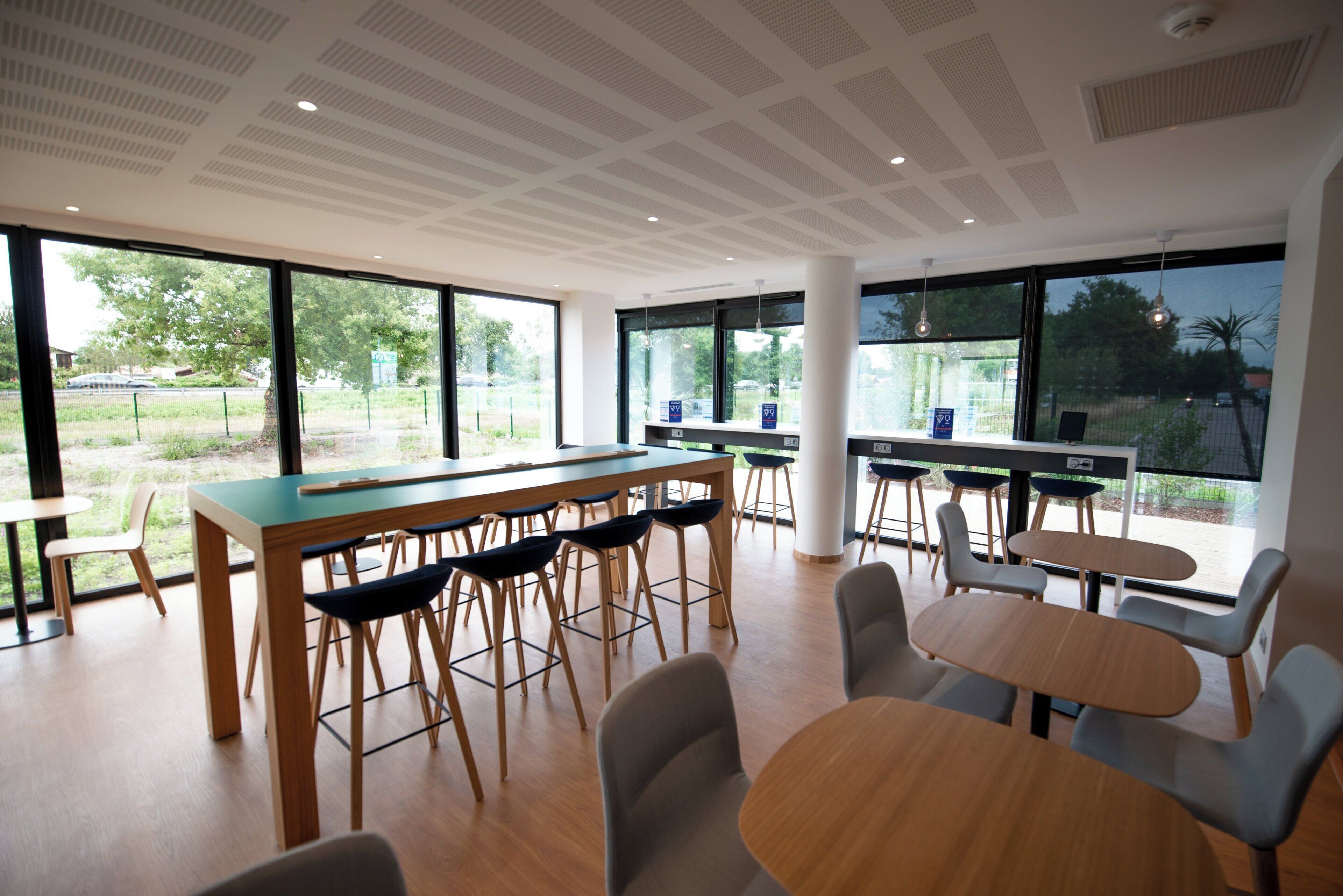 Un espace intérieur moderne doté d’une longue table et de nombreuses chaises, de grandes fenêtres et d’un plafond orné d’un motif en réseau.