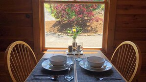 A wooden dining table set for two with plates, cutlery, and glasses, positioned in front of a window with a view of trees and a garden. - A view for 2 & Cozy, Close to PF and Gatlinburg (Gatlinburg)