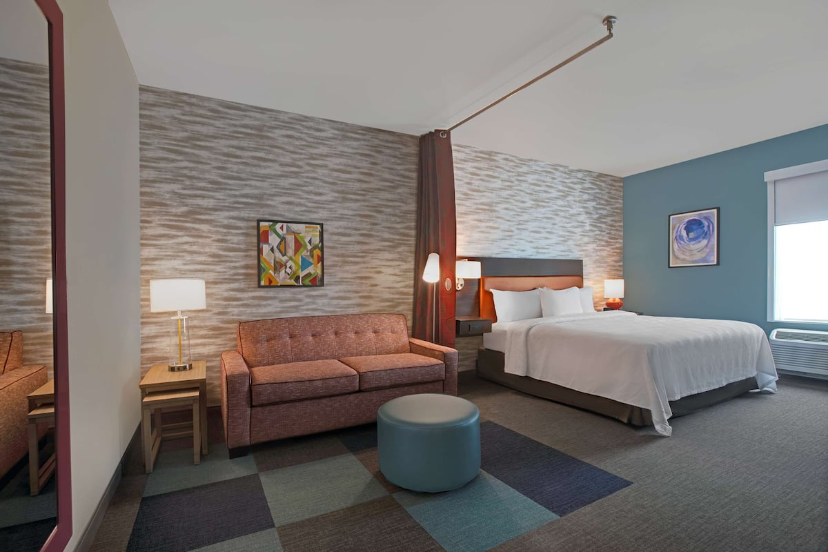 A hotel room with a bed, a sofa, a round ottoman, a nightstand with a lamp, and a wall with a textured pattern.
