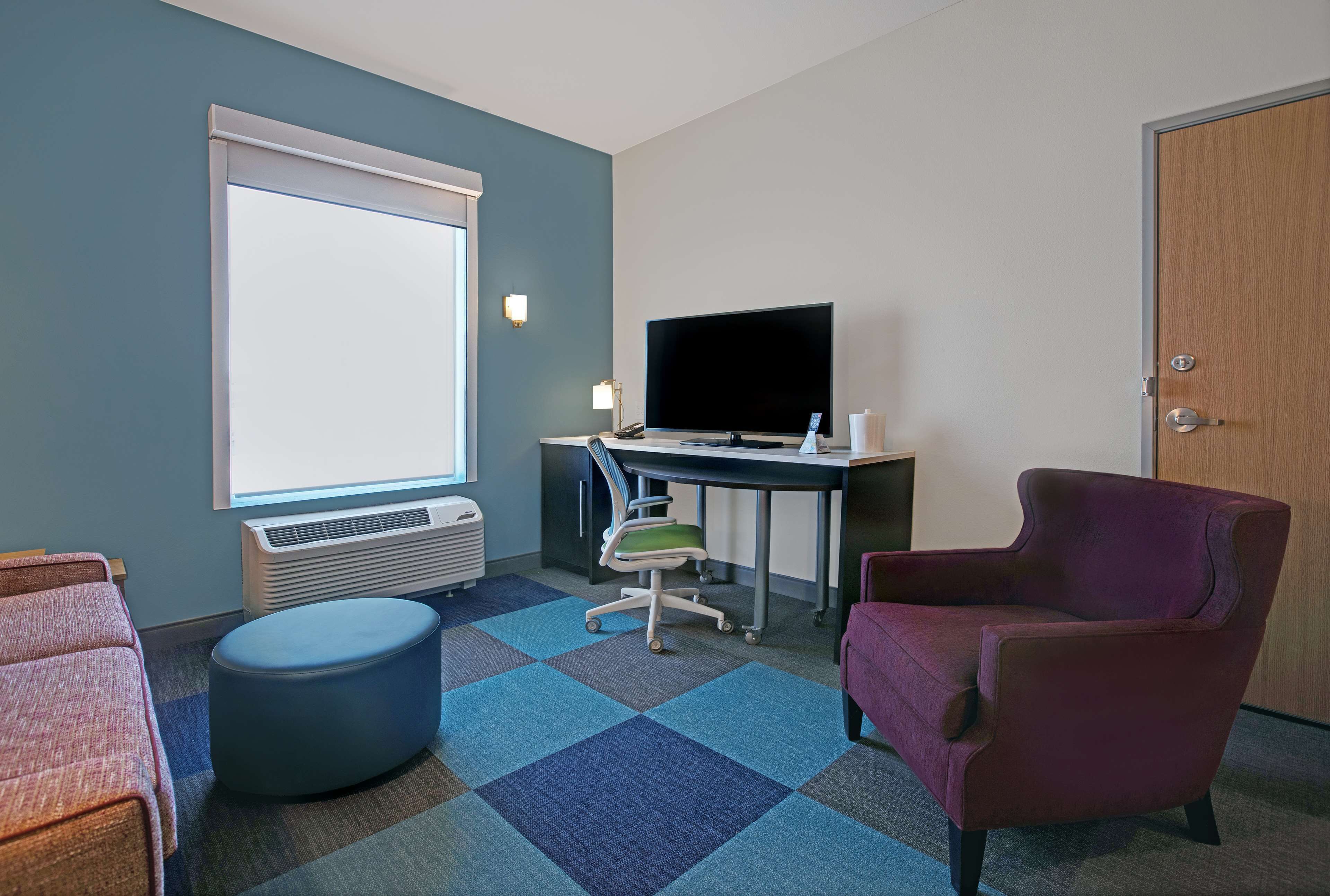 A hotel room with a large window, a flat-screen TV, a desk, a purple armchair, a blue ottoman, and a red sofa.