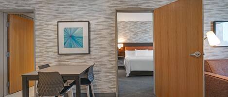 Una habitación de hotel con cama, un escritorio con sillas, un cuadro en la pared y una puerta que da a otra habitación.