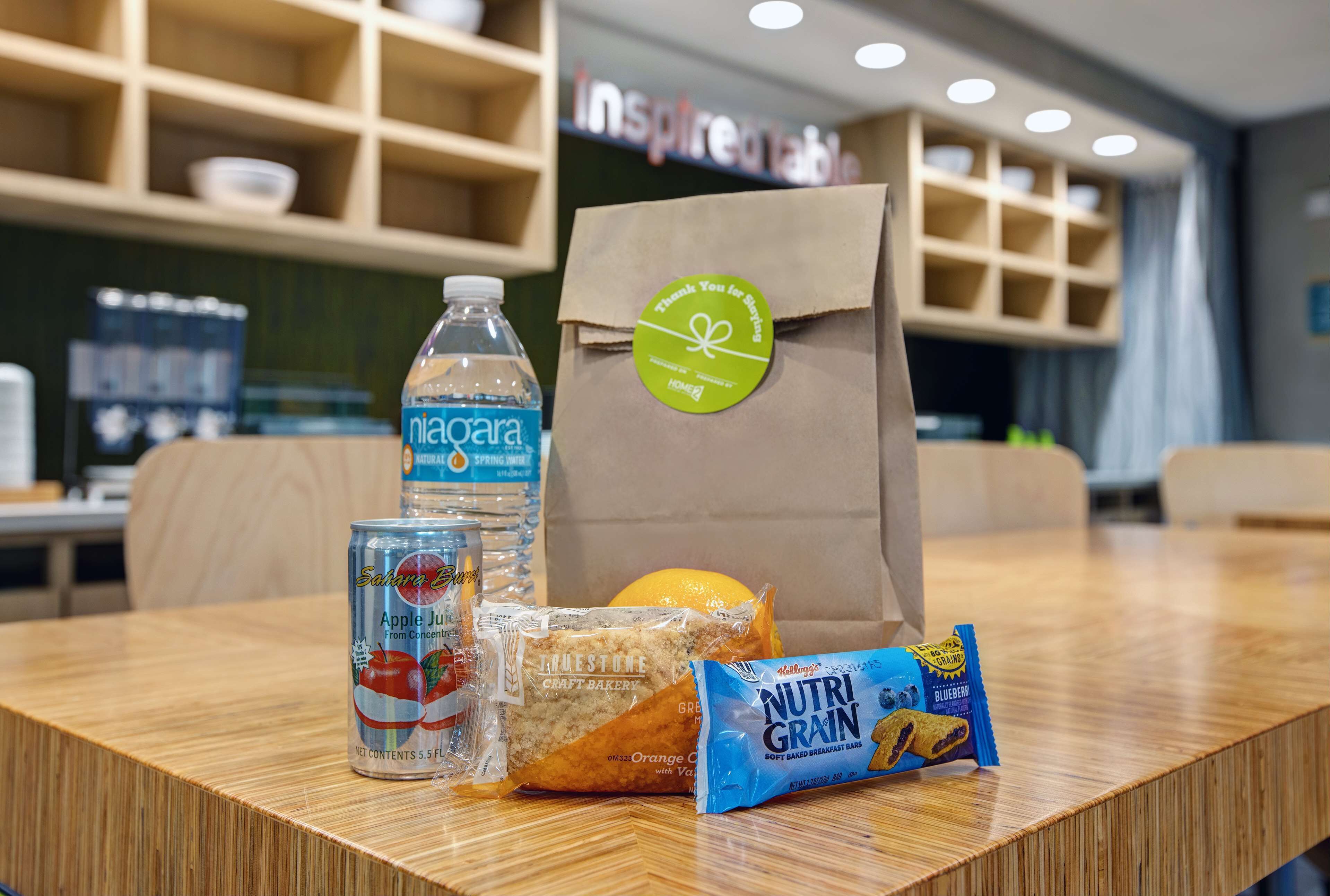 A table with a bottle of water, a can of apple juice, a loaf of bread, and a bag of Nutri Grain bars.
