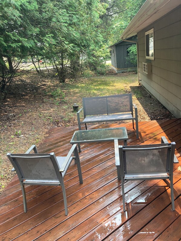 An outdoor deck with a bench and chairs, surrounded by trees and a house.