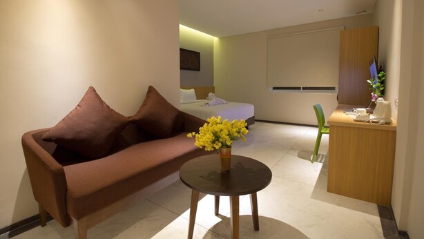 A modern hotel room with a brown sofa, a small round table with a vase of yellow flowers, a wooden desk, and a bed in the background. - Vinotel Cirebon (West Cirebon)