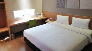 A hotel room with a bed, a desk, a chair, a small window, and a trash can. - Vinotel Cirebon (West Cirebon)