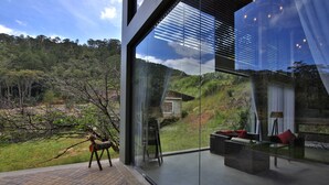 Una habitación moderna con paredes de vidrio y vistas a un paisaje exuberante y una casa pequeña a lo lejos.