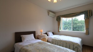 A room with two beds, a small table with a lamp, an air conditioning unit, and a window with curtains. - Pension Tinkuna (Minamiaso)