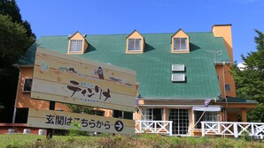 A building with a green roof and orange walls, featuring a large sign with Japanese text and a directional arrow. - Pension Tinkuna (Minamiaso)