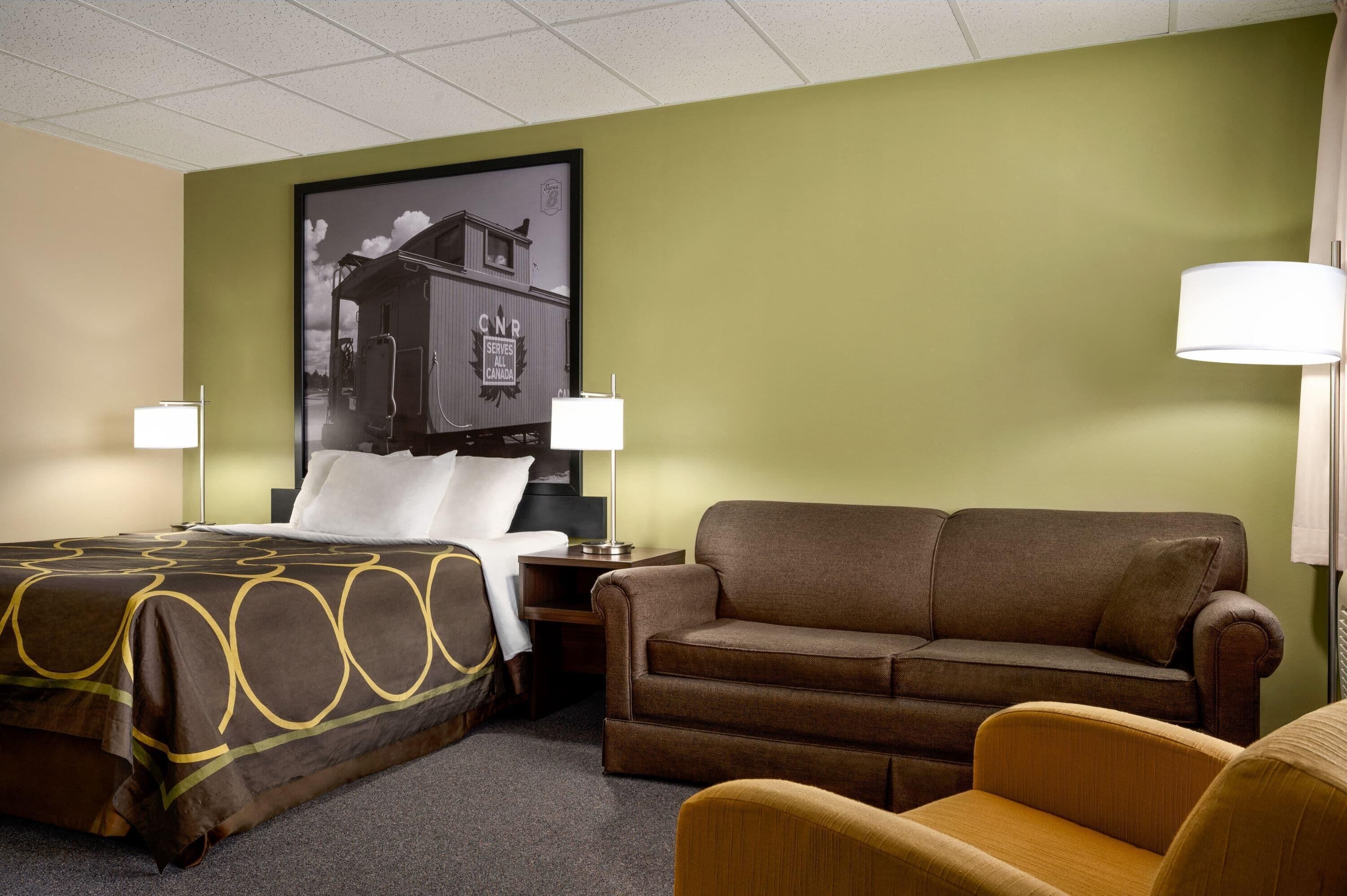 A hotel room with a bed, a brown sofa, a small table with a lamp, and a poster on the wall.