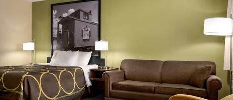 A hotel room with a bed, a brown sofa, a small table with a lamp, and a poster on the wall.