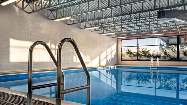 Una piscina interior con escaleras metálicas, pared de ladrillo y ventanas amplias con vistas a árboles.