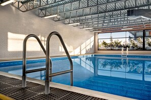 An indoor swimming pool with metal ladders, a brick wall, and large windows with a view of trees. - Super 8 by Wyndham Hearst ON (Hearst)