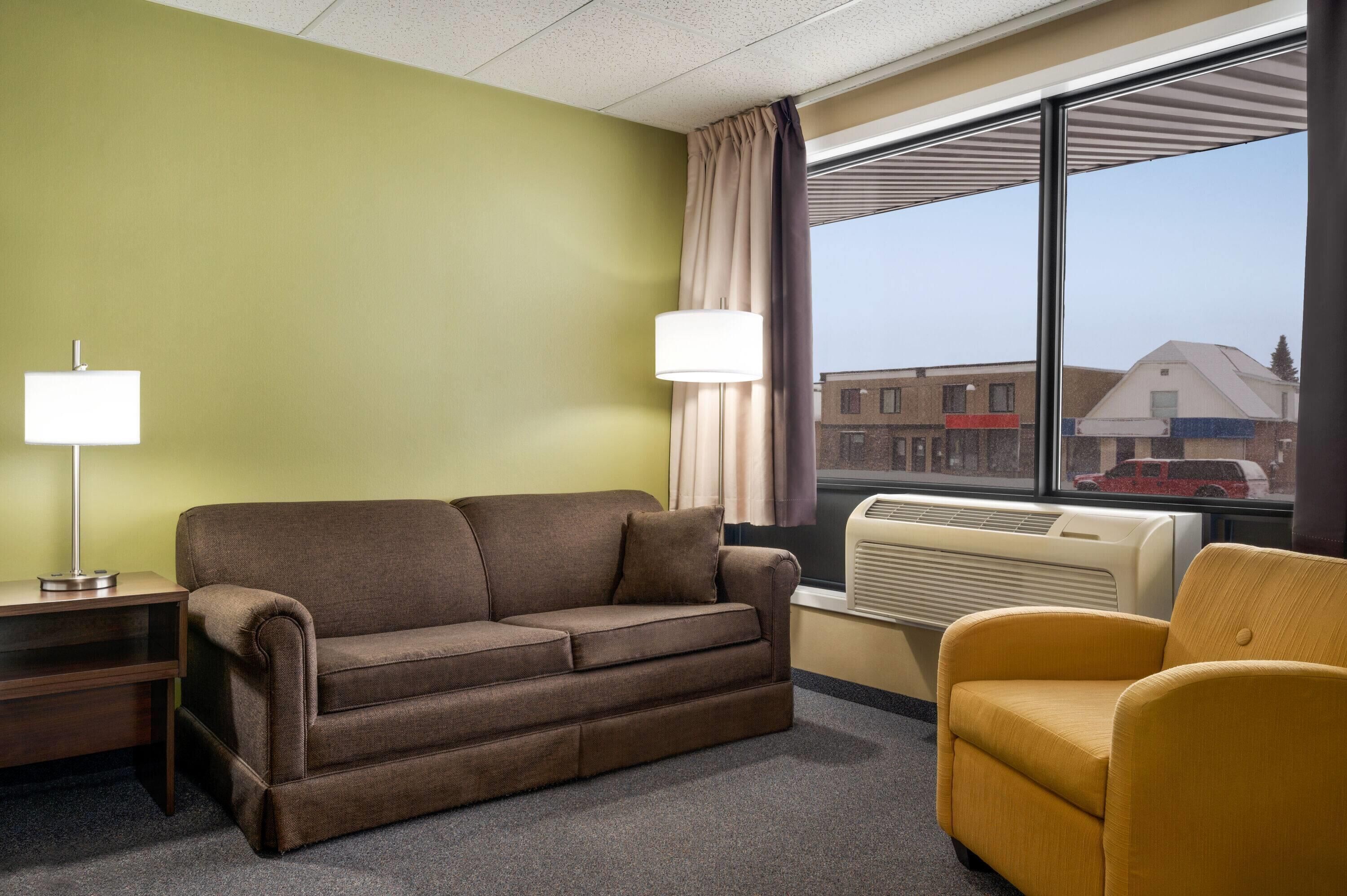 A hotel room with a brown sofa, a yellow armchair, a lamp, and a window with a view of buildings and a red car outside.