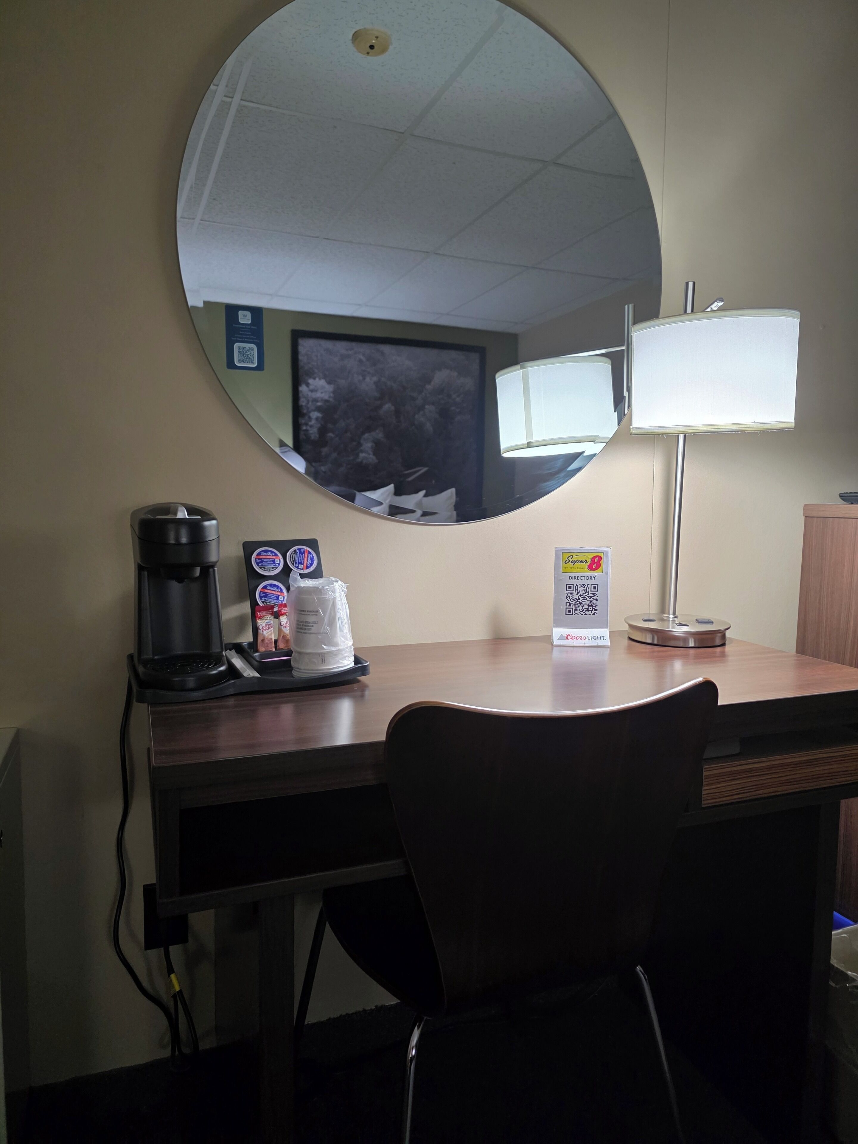 Ein Hotelzimmer mit einem runden Spiegel, einem Schreibtisch mit Kaffeemaschine, einer Lampe und einem Sessel.