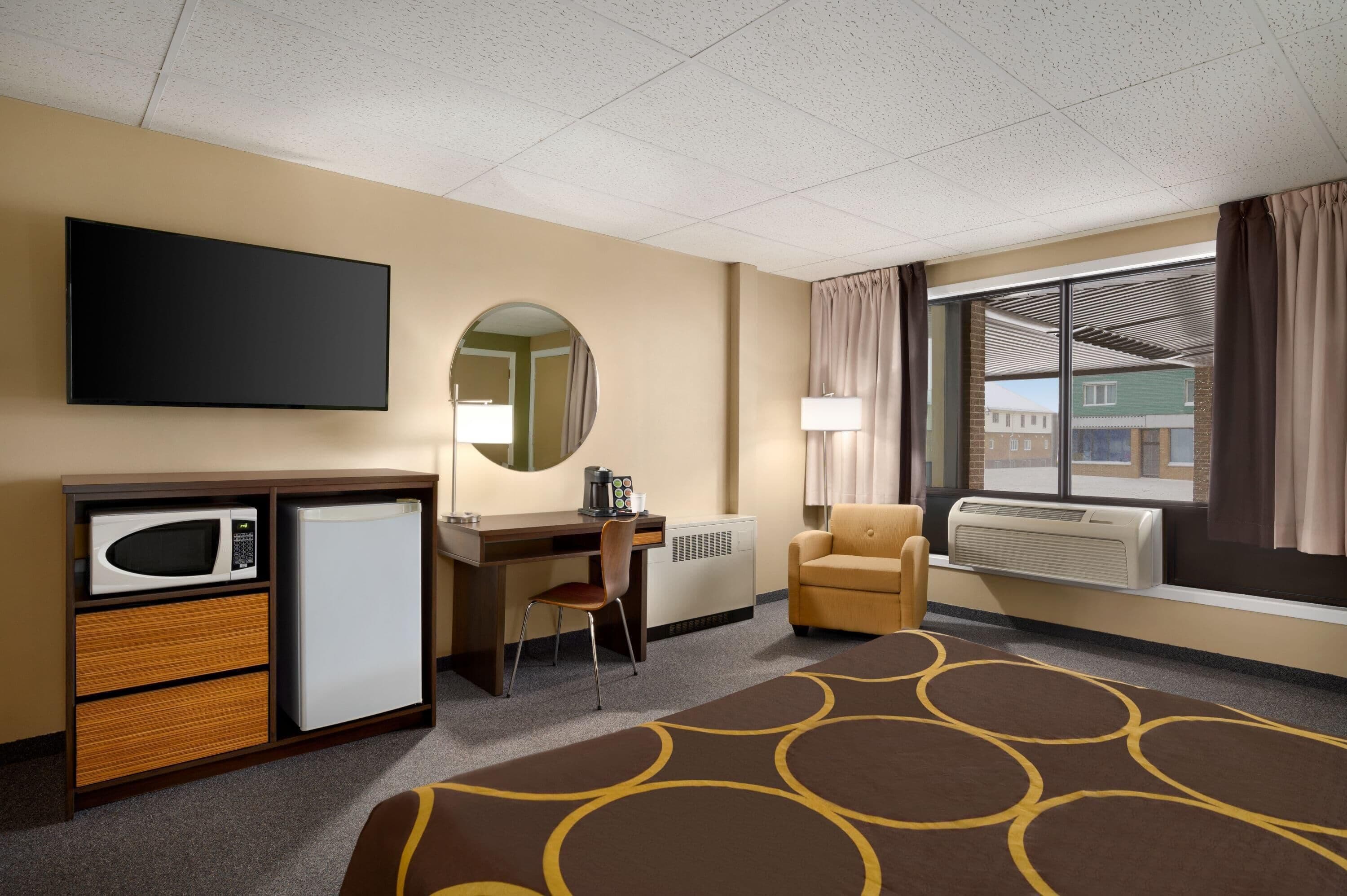 A hotel room with a bed, a desk, a chair, a microwave, a refrigerator, a TV, a window with curtains, and a radiator.
