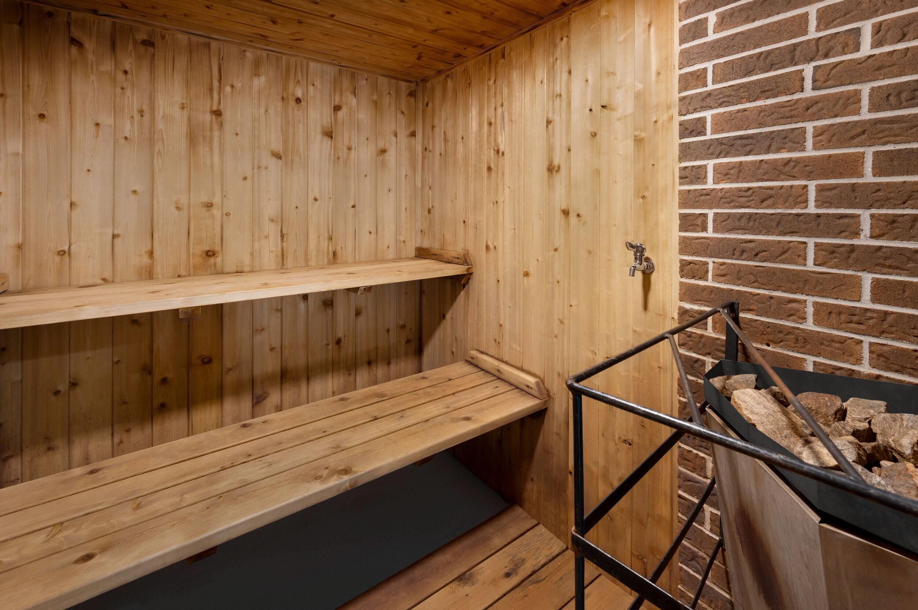 A sauna with wooden walls and a brick wall section. There are benches and a metal rack with logs.