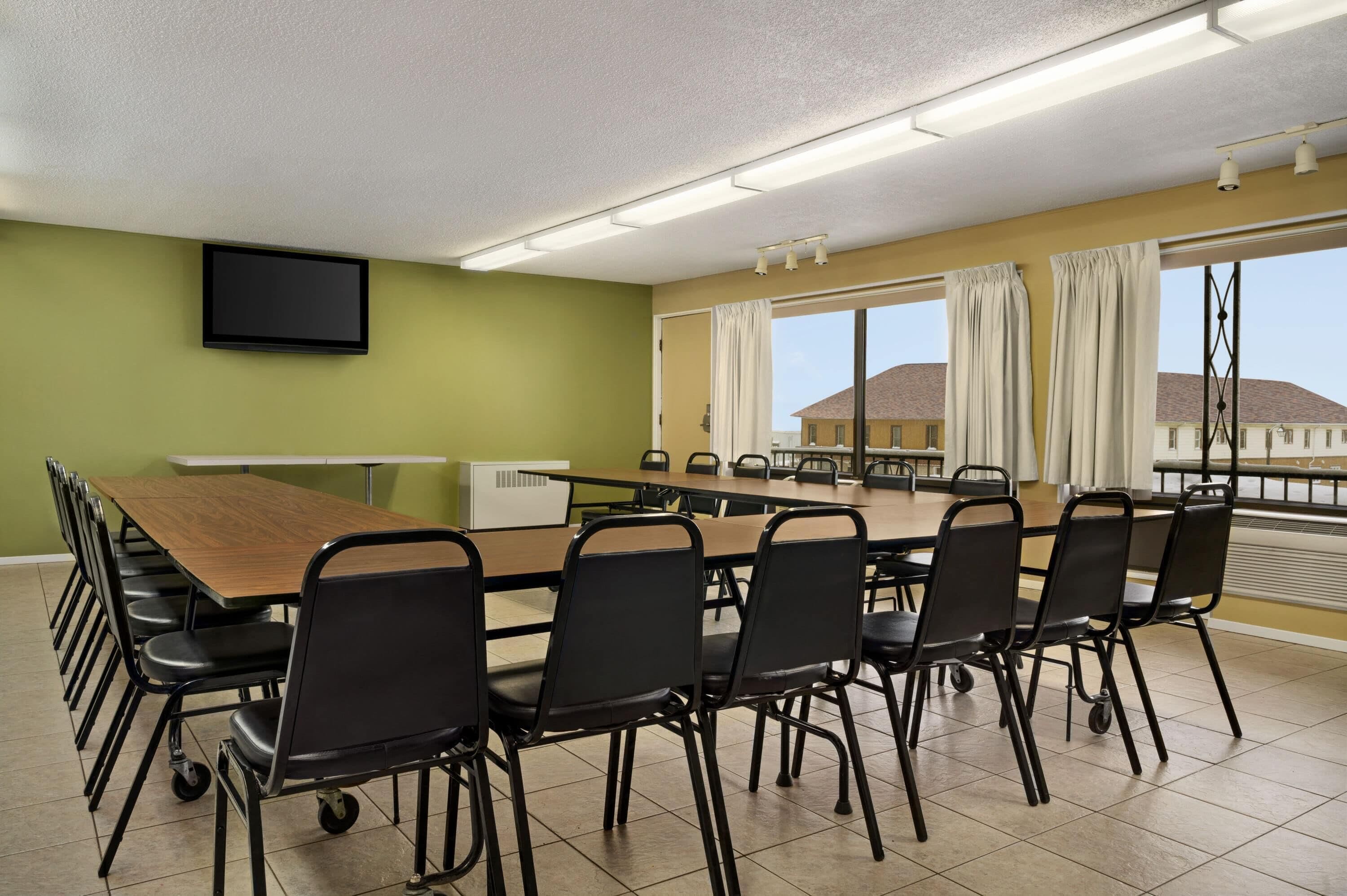 Un salón de conferencias con una larga mesa y sillas, un televisor de pantalla plana en la pared y amplias ventanas con cortinas.