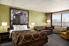 A hotel room with a large bed, a sofa, a chair, a window with a view, and a wall-mounted air conditioner.