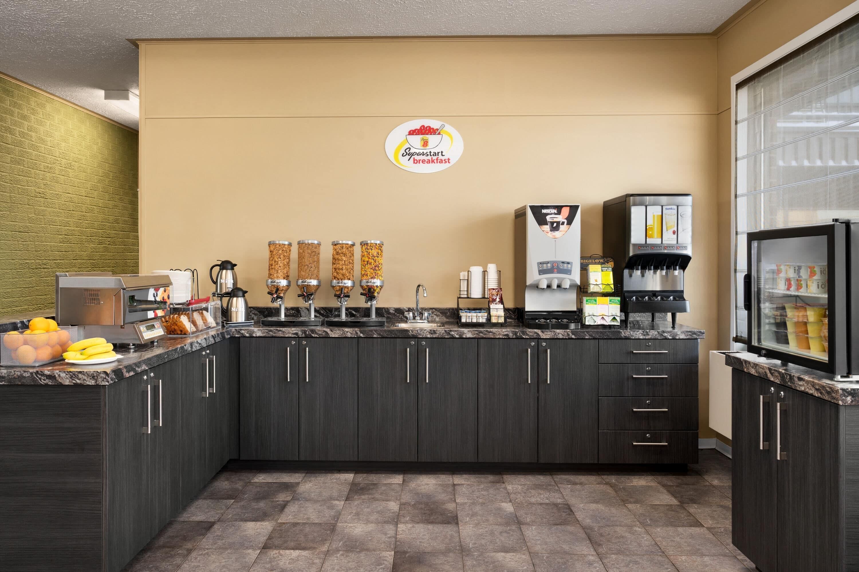 Una barra de desayuno con una máquina de café, dispensadores de jugo y un refrigerador.