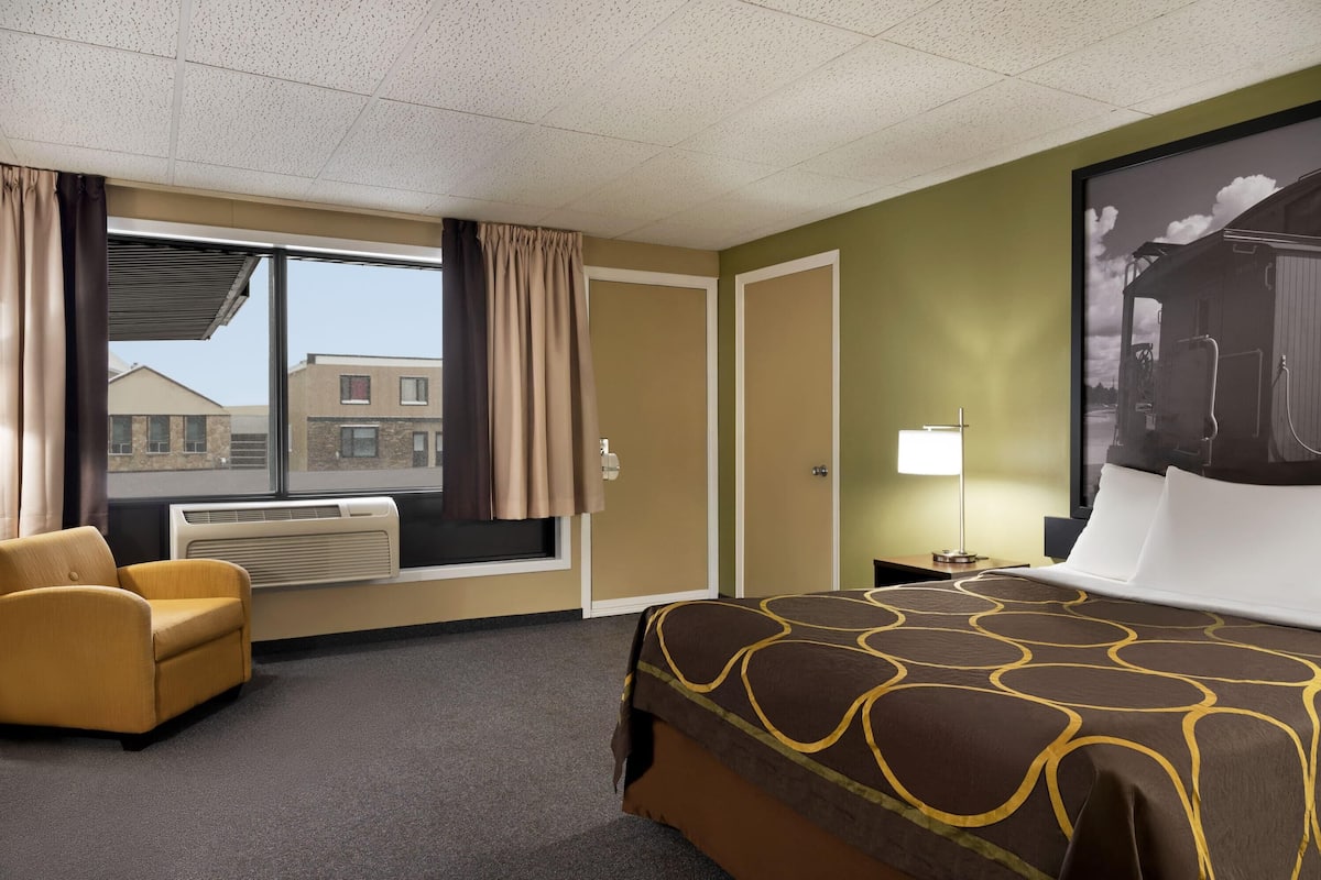 Een hotelkamer met een bed, een stoel, een raam met gordijnen, een radiator en een wandlamp.