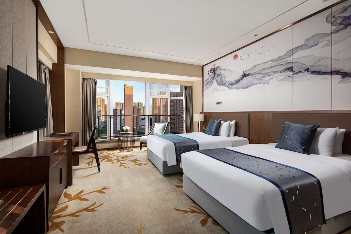 A hotel room with two beds, a desk, a TV, and a large window with a city view.