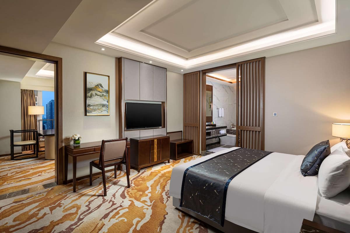 A hotel room with a large bed, a desk with a chair, a flat-screen TV, and a view of the cityscape.